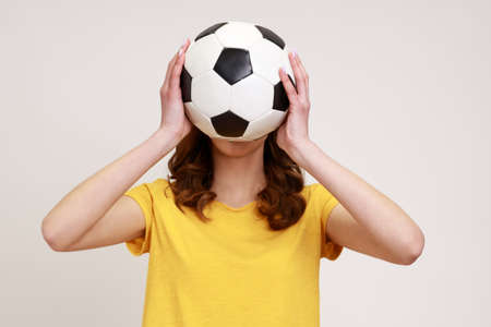 Unknown female with wavy hair football fan in casual style yellow t-shirt supporting her favorite team covering face with soccer ball. Indoor studio shot isolated on gray background.の写真素材