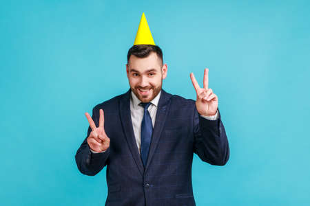 Happy bearded guy with funny party cone on head showing victory sign with both hands, looking at camera with toothy smile, celebrating birthday holiday. Indoor studio shot isolated on blue background.の写真素材