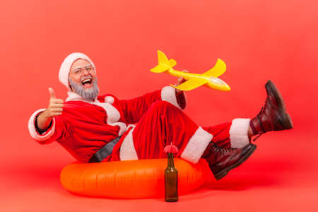 Happy elderly man wearing santa claus costume sitting on orange rubber ring and holding yellow...