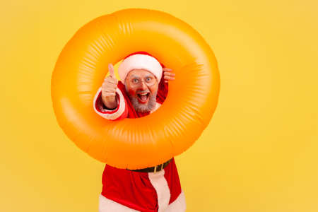Extremely happy elderly man with gray beard wearing santa claus costume posing with orange rubber ring and showing thumb up, enjoying winter vacation. Indoor studio shot isolated on yellow background.の写真素材
