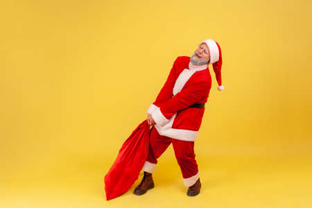 Elderly man with gray beard wearing santa claus costume dragging his very large and heavy bag full of presents, frowning face, trying to lift up. Indoor studio shot isolated on yellow background.の写真素材