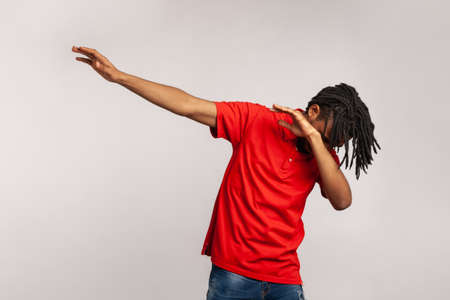 Portrait of young adult man with dreadlocks wearing red casual style T-shirt, standing in dab dance pose, internet meme, celebrating success. Indoor studio shot isolated on gray background.の写真素材