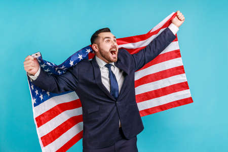 Happy businessman in suit raising American flag, shouting for joy celebrating US Independence day 4th of july, government business support. Indoor studio shot isolated on blue background.の写真素材