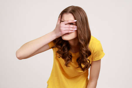 Portrait of beautiful teenager girl wearing yellow casual T-shirt spying, hiding and peeping through fingers, looking for secrets, curiosity. Indoor studio shot isolated on gray background.の写真素材