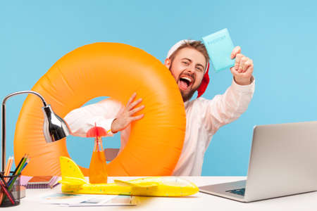 Excited happy man office worker in santa claus hat sitting with toy plane, lemonade and in rubber ring, showing passport, going on winter vacation. Indoor studio shot isolated on blue backgroundの写真素材