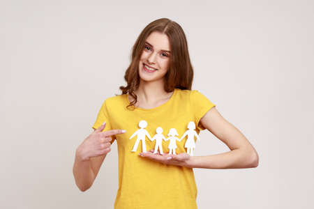 Kind smiling positive teenager girl in yellow t-shirt pointing at paper chain people which holding in hands, happy family, relationships, parenthood. Indoor studio shot isolated on gray background.の写真素材