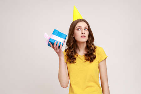 Curious adorable woman of young age in yellow casual T-shirt shaking blue giftbox near ear trying to...