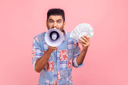 Mad bearded young adult man wearing blue casual style shirt screaming in megaphone holding fan of dollars, announcing bonuses and promotions. Indoor studio shirt isolated on pink background.の写真素材