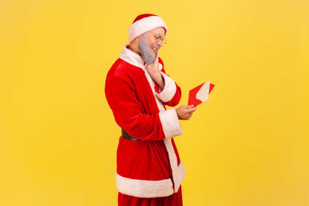 Profile portrait of elderly man with gray beard wearing santa claus costume being touched with sensitive letter, reading Christmas congratulations. Indoor studio shot isolated on yellow background.の写真素材