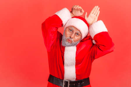 Funny elderly man with gray beard wearing santa claus costume pout lips playfully and showing bunny ears, aping with comical childish grimace. Indoor studio shot isolated on red background.の写真素材