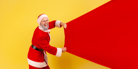 Side view portrait of elderly man with gray beard wearing santa claus costume carrying big bag with presents, copy space for advertisement. Indoor studio shot isolated on yellow background.の写真素材