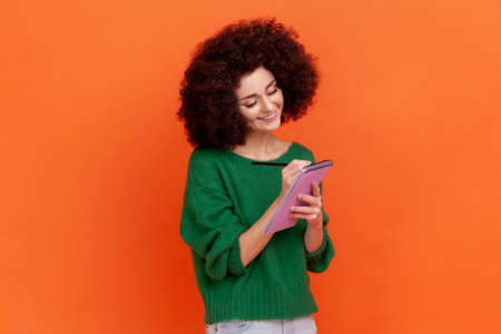 Positive smiling woman with curly hair wearing green casual style sweater writing down in paper...