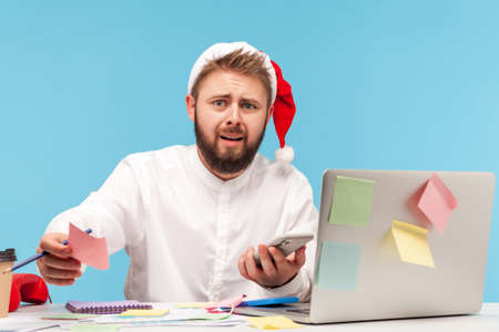 Unhappy anxious man in santa claus hat sitting at workplace all covered with sticky notes, writing down information holding smartphone in hand. Indoor studio shot isolated on blue backgroundの写真素材