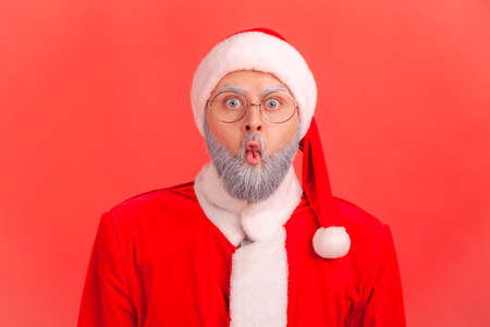 Elderly man with gray beard wearing santa claus costume making fish face with pout lips and surprised shocked expression, looking with big eyes. Indoor studio shot isolated on red background.の写真素材
