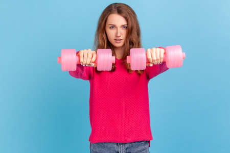 Winsome confident woman with wavy hair wearing pink pullover standing holding out two dumbbells,...