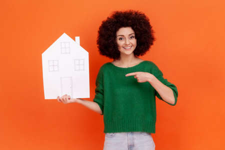 Portrait of woman with curly hair wearing green casual style sweater pointing at paper house in her hand, looking at camera with toothy smile. Indoor studio shot isolated on orange background.の写真素材