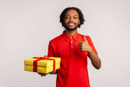 Bearded man with toothy smile wearing red casual style T-shirt, standing and holding yellow box and...