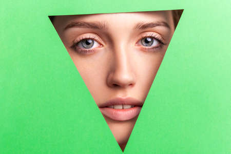 Closeup portrait of well-groomed woman with natural makeup, looking at camera through triangular hole of green paper, beauty and health and skin care concept. Indoor studio shot.の写真素材