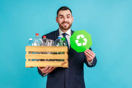 Satisfied man wearing official style suit holding box with plastic bottles and recycling green symbol, sorting his rubbish, saving ecology. Indoor studio shot isolated on blue background.の写真素材