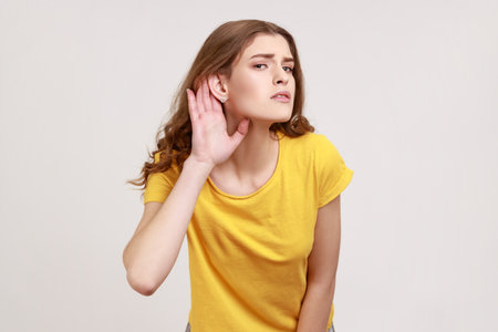 What? I can't hear! Portrait of girl in yellow T-shirt holding hand near ear and listening...