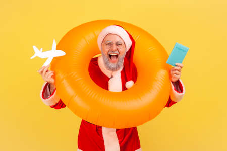 Excited elderly man with gray beard in santa claus costume standing with rubber ring over neck, holding paper plane and passport in hands, winter tour. Indoor studio shot isolated on yellow backgroundの写真素材