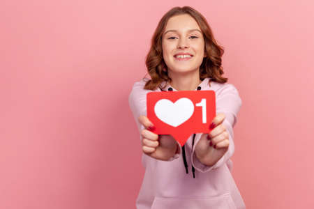 Portrait of cheerful smiling teenage girl showing heart icon at camera, recommending to follow, like me sign. Indoor studio shot isolated on pink backgroundの写真素材