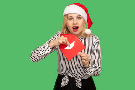 Portrait of excited blonde young woman in christmas hat standing with surprised face and opening christmas greeting envelope, looking with amazed face. indoor studio shot, isolated on green backgroundの写真素材
