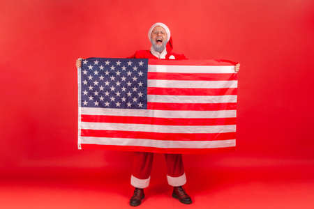 Yelling elderly man with gray beard wearing santa claus costume showing USA flag and screaming, celebrating enjoying Christmas holidays. Indoor studio shot isolated on red background.の写真素材