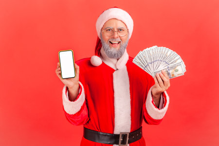 Happy smiling elderly man with gray beard wearing santa claus costume holding fan of money and showing smartphone with white blank screen. Indoor studio shot isolated on red background.の写真素材