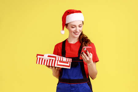 Positive delivery woman holding present box and using smart phone, specifies delivery address,...
