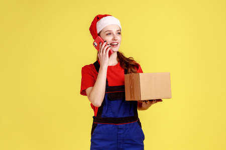 Friendly delivery woman talking on cellphone with client and accepting order, holding cardboard box, wearing blue overalls and santa claus hat. Indoor studio shot isolated on yellow background.の写真素材