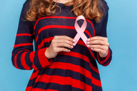 Anonymous woman wearing striped casual style sweater, woman holding pink ribbon, symbol of breast cancer awareness, female healthcare. Indoor studio shot isolated on blue background.の写真素材