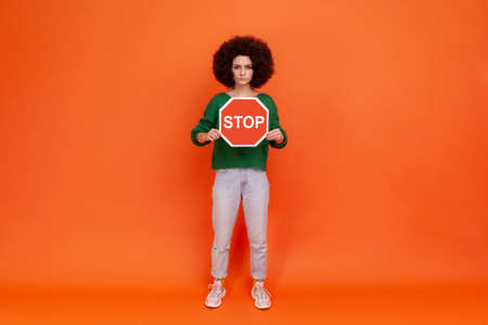 Full length portrait of strict woman with Afro hairstyle wearing green sweater standing with serious...