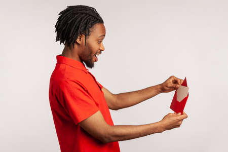 Side view of positive man with dreadlocks wearing red casual style T-shirt, opening letter in red envelope, holding greeting card and feels happy. Indoor studio shot isolated on gray background.の写真素材