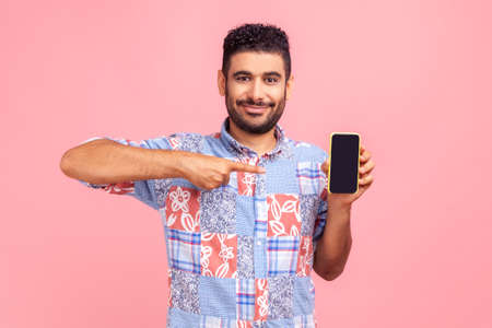 Happy cheerful beaded man in blue casual shirt shirt pointing finger at smartphone with empty screen, looking at camera with smile, freespace for adv. Indoor studio shot isolated on pink background.の写真素材