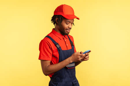 Side view portrait of positive deliveryman wearing uniform, using smartphone for taking new order for delivering, delivery service. Indoor studio shot isolated on yellow background.の写真素材