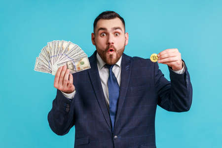 Portrait of young adult bearded man wearing official style suit with astonished facial expression showing to camera dollars banknotes and golden bitcoin. Indoor studio shot isolated on blue backgroundの写真素材
