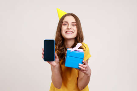 Birthday present, bonus for mobile user. Portrait of happy teenager girl in birhday cone holding gift box and cell phone with mock up, blank display. Indoor studio shot isolated on gray background.の写真素材