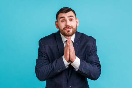 Please, I'm begging! Unhappy bearded man in official style suit keeping hands in prayer looking with imploring expression, sincere asking permission. Indoor studio shot isolated on blue background.の写真素材