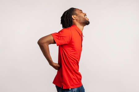 Side view of guy with dreadlocks wearing red casual style T-shirt, holding his back, having sudden...