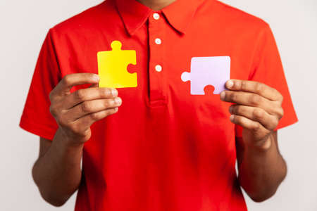 Faceless person wearing red casual style T-shirt, holding two puzzle parts, connecting colorful jigsaw pieces, symbol of unity and association. Indoor studio shot isolated on gray background.の写真素材