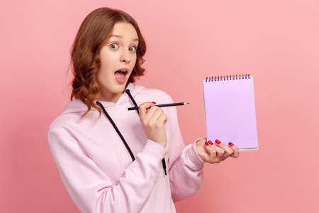 Portrait of surprised curly haired teenage girl in hoodie with opened mouth pointing pencil at empty notebook sheet, place for advertisement. Indoor studio shot isolated on pink backgroundの写真素材