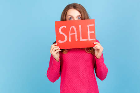 Portrait of curious woman wearing pink pullover, peeping from red card with sale inscription, mega shopping, black friday discounts. Indoor studio shot isolated on blue background.の写真素材