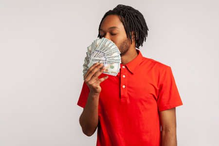 Profile of young adult guy in red casual T-shirt, smelling earned dollar banknotes, enjoying success and big profit, wealthy life, greedy for money. Indoor studio shot isolated on gray background.の写真素材