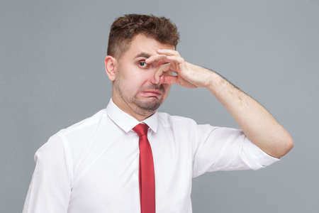 bad smell. Portrait of young confused man in white shirt and tie standing pinching his nose and looking at camera because back smell. indoor isolated on gray background.の写真素材
