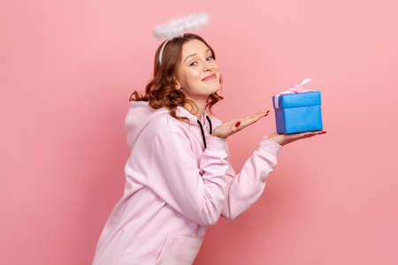 Side view portrait of smiling curly haired teenage girl in hoodie and halo on head holding blue wrapped gift box, holiday greeting. Indoor studio shot isolated on pink backgroundの写真素材