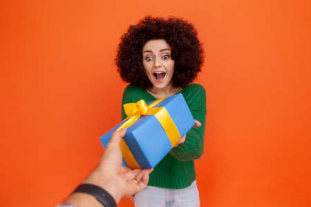 Portrait of happy excited woman with Afro hairstyle wearing green casual style sweater getting blue wrapped present box from unknown person. Indoor studio shot isolated on orange background.の写真素材