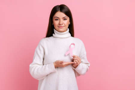 Portrait of calm beautiful woman standing holding rose ribbon in front of chest, female healthy, wearing white casual style sweater. Indoor studio shot isolated on pink background.の写真素材