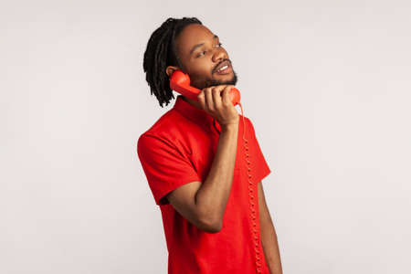 Side view of male with dreadlocks and beard wearing red casual style T-shirt, making calls on landline telephone, looking at camera with toothy smile. Indoor studio shot isolated on gray background.の写真素材