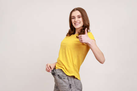 Profile portrait of beautiful happy teenager girl have willpower showing slim waist in big trousers and showing thumbs up, successful weight loss. Indoor studio shot isolated on gray background.の写真素材
